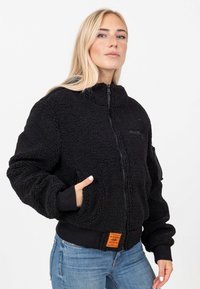Bomber jacket nera realizzata in materiale morbido e testurizzato, dotata di chiusura con zip, tasche laterali e un'etichetta arancione in vita.