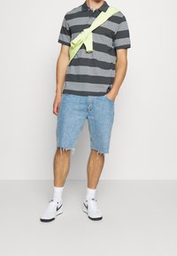 Muž oblečený v šedém a černém pruhovaném polo tričku, s světle zeleným svetr přehodeným přes ramena, v světle modrých denimových šortkách, s bílými ponožkami a bílými teniskami Nike.