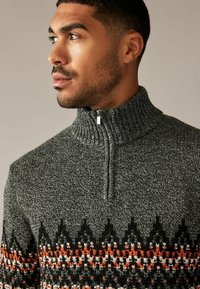 Pull en tricot gris avec un col montant, présentant un motif noir, blanc et orange près de l'ourlet et une fermeture éclair sur le devant à mi-hauteur.