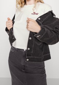 Černá džínová bunda se stříbrnými knoflíky, nošená přes bílé tričko s červeným výšivkou "Levi's", doplněná o černou sukni ve stejném odstínu.