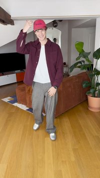 Vīna sarkana pogu kreklā, balts T-krekls, pelēkas teksturētas bikses un sudraba sporta apavi. Valkā sarkanu cepuri ar tekstu. Iekštelpu vide ar zaļumiem.