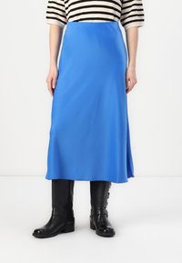 Longue jupe bleu en tissu lisse, présentant une silhouette droite. Associée à des bottes en cuir noir montant jusqu'aux genoux avec des boucles.