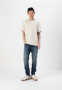 Beige kortærmet t-shirt, blå slim-fit jeans og hvide sneakers med brune såler, stående mod en ensfarvet hvid baggrund.