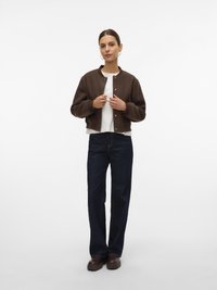 Femme portant une bomber jacket marron à boutons, un haut blanc, un jean bleu foncé et des chaussures marron, se tenant devant un fond blanc.