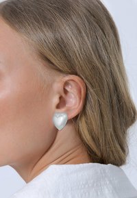 Herzförmige Ohrringe in einem glänzenden metallischen Silberfinish, mit einer glatten Oberfläche und einer klaren Ohrstecker, getragen am Ohr eines Models.