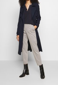 Marineblauer Trenchcoat über einer dunklen Bluse, kombiniert mit hellgrauen, gemusterten Hosen und schwarzen Stiefeletten mit quadrater Spitze und Blockabsatz.