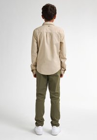 Kind dat een lichtbeige overhemd met lange mouwen, groene broek en witte sneakers draagt, gezien vanaf de achterkant.