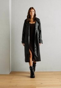 Manteau trench en vinyle noir avec de grands revers, des boutons et une fente jusqu'à la cuisse, associé à des bottines lacées noires, sur fond neutre.