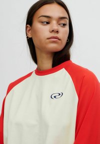 Młoda kobieta z długimi ciemnymi włosami, ubrana w biało-czerwony t-shirt raglanowy z małym granatowym logo w kształcie spirali, patrząca lekko w lewo.