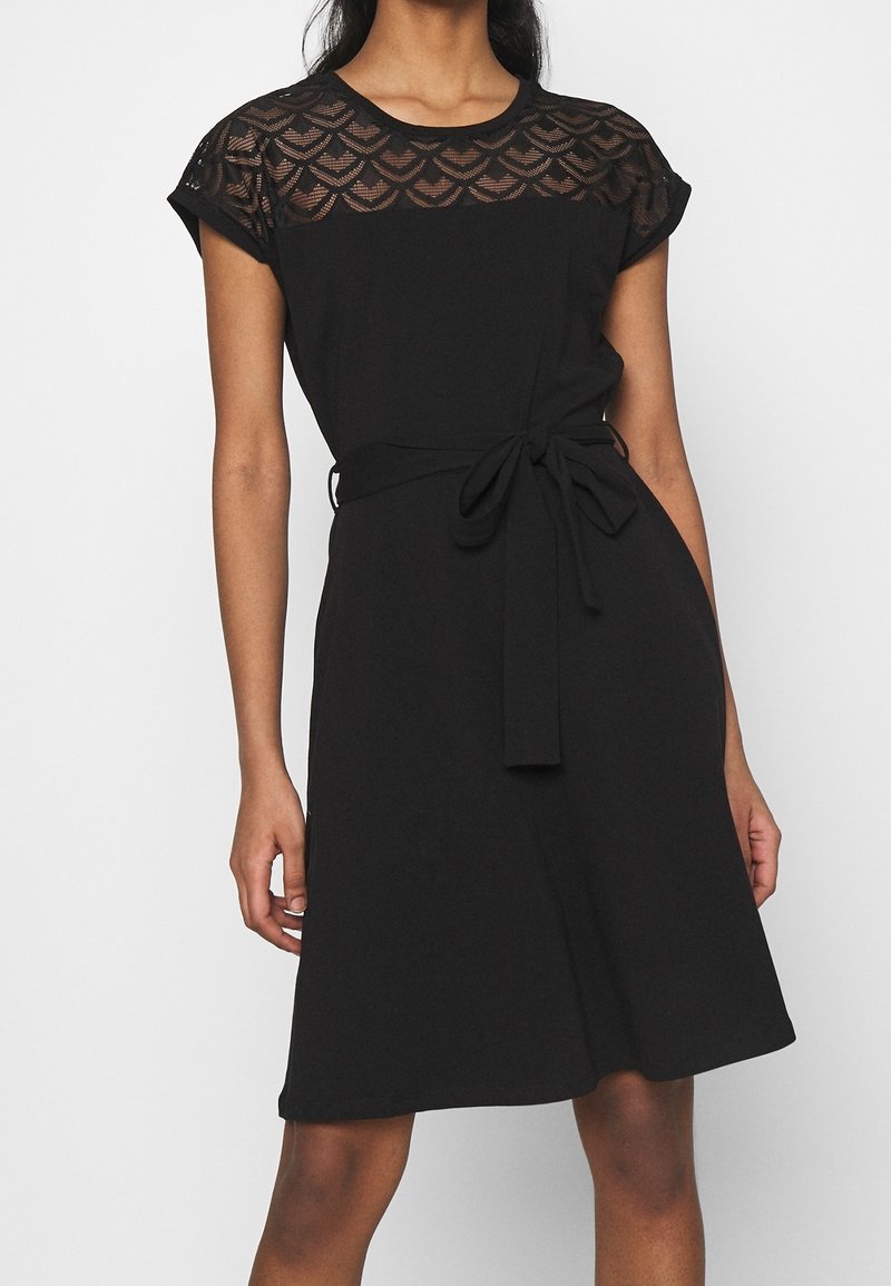 Femme portant une robe noire à la longueur genou avec un motif en dentelle sur la partie supérieure de la poitrine et les manches, ceinture en tissu nouée à la taille.