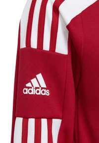 Rød atletisk trøje med hvidt tre-stripes design på ærmerne, tekstureret stof og et hvidt Adidas-logo på overarmen.