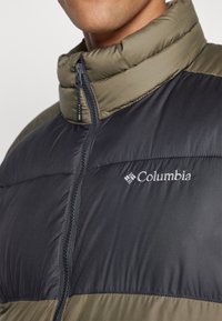Melna un olīvu zaļa pūkainā jaka ar augstu apkakli, rāvējslēdzēja aizdari un Columbia logo uz krūtīm. Gluda, izolēta auduma tekstūra.
