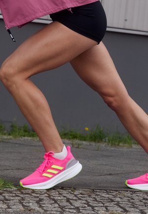 Personne portant des chaussures de course rose fluo et un short noir en pleine foulée sur une surface extérieure pavée.