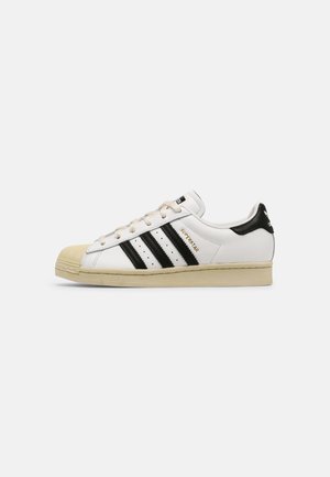 Weißer Adidas Superstar Sneaker mit schwarzen Streifen, beiger Sohle und Zehenschutz sowie goldenem "Superstar" Text an der Seite.