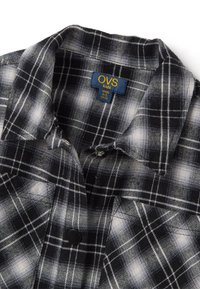 Camicia di flanella a scacchi bianca e nera con colletto, chiusura a bottoni e un'etichetta che mostra "OVS kids" e informazioni sulla taglia.