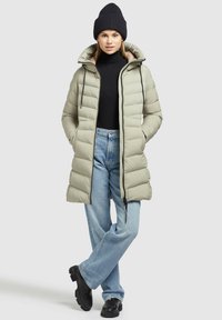 Kvinde iført en beige pufferjakke, sort beanie, sort turtleneck, lyseblå jeans og sorte støvler, stående med hænderne i lommerne.