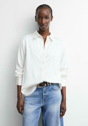 Femme aux cheveux courts tressés portant une chemise blanche boutonnée rentrée dans un jean bleu avec une ceinture noire, debout contre un mur blanc.