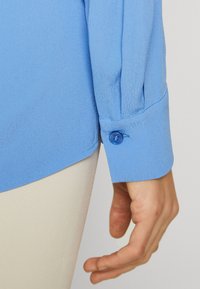 Camicia azzurra chiaro a maniche lunghe con polsino abbottonato, texture liscia e orlo leggermente curvo. Mani visibili al polsino.