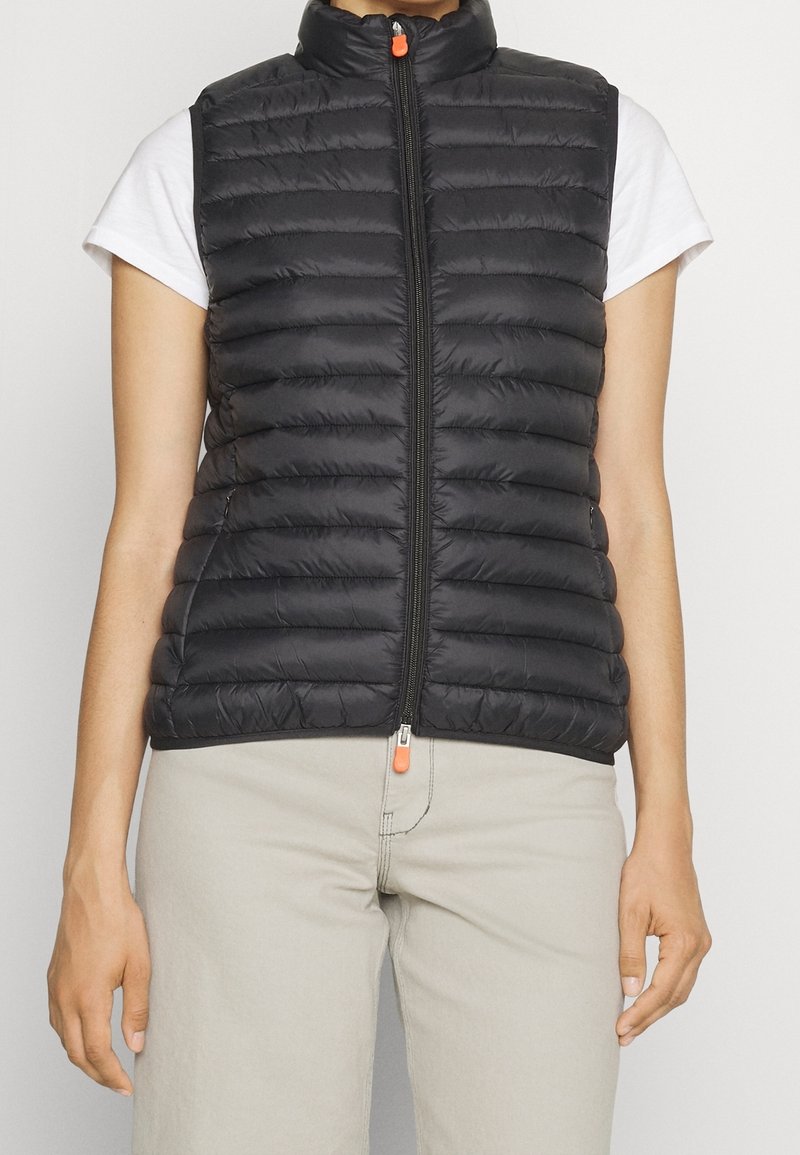 Chaleco negro acolchado con cuello alto, diseño acolchado y cremallera frontal. Presenta un tirador de cremallera en contraste de color naranja. Lleva puesto sobre una camisa blanca.