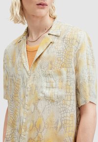 Camicia a maniche corte in un tessuto leggero e fantasia con stampa di serpente gialla e grigia. Include una tasca sul petto e chiusura con bottoni.