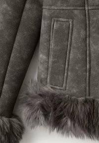 Mangas de chaqueta de ante gris oscuro con detalle de costura rectangular y grueso ribete de piel gris en los puños sobre un fondo claro.