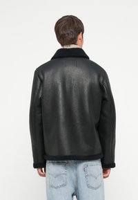 Schwarze Lederjacke mit einem Schaffellkragen und -manschetten, mit einer strukturierten Oberfläche und kastiger Form, getragen über hellblauen Jeans.