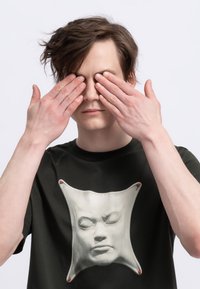 Zwarte T-shirt met een wit, gesculpteerd gezicht ontwerp in het midden. Het gezicht heeft verhoogde kenmerken en is geplaatst tegen een gladde stofachtergrond.