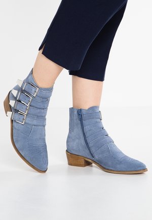 Bottines en daim bleu clair avec petits talons en bois, plusieurs brides avec boucles d'un côté et une fermeture éclair de l'autre, portées avec un pantalon court foncé.