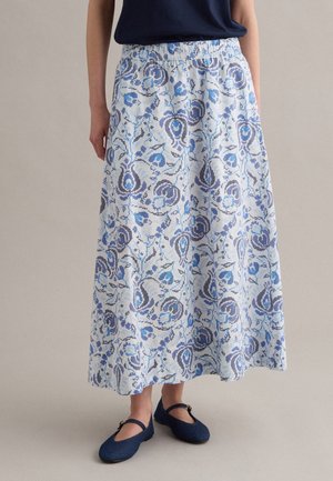 Donna con gonna midi bianca con motivi floreali blu, top blu navy e scarpe Mary Jane blu navy, in piedi su sfondo neutro.