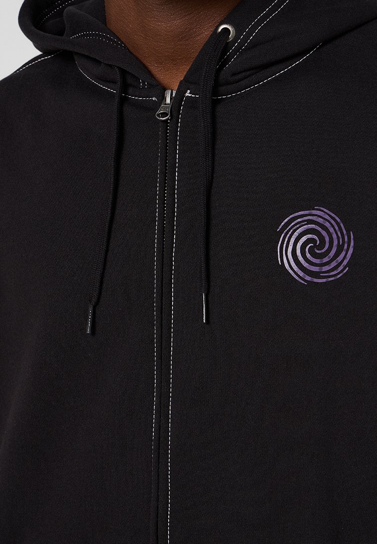 Sudadera negra con cremallera y costuras blancas, y un diseño en espiral morado en el lado izquierdo del pecho, usada por una persona de piel morena.
