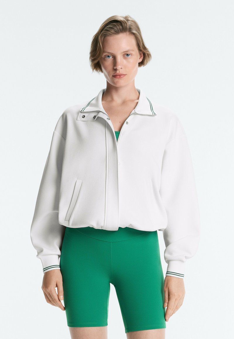 Jeune femme aux cheveux blonds courts portant une veste blanche zippée et un short de cycliste vert taille haute, debout devant un fond uni.