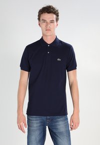 Temno moderna polo majica iz bombaža, z ovratnikom, dvema gumboma in majhnim zelenim logotipom na levi prsni strani. Ima kratke rokave.
