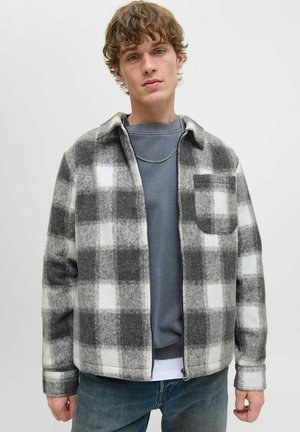 Young man wearing a black, white, and gray checkered zip-up jacket over a gray sweatshirt and blue jeans against a plain light background.