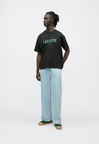 T-shirt preta de algodão com logótipo "LACOSTE Paris" verde e roxo, combinada com jeans de perna larga azul-claro e ténis verdes.