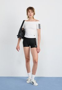 Top branco com ombros à mostra com riscas pretas, combinado com calções pretos com riscas a condizer, finalizado com meias brancas e ténis.