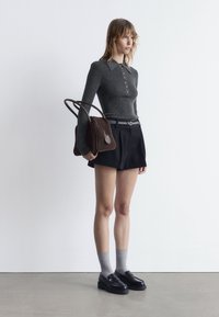 Graues, geripptes Langarmoberteil mit Knöpfen, schwarze Shorts, graue Socken und schwarze Slipper. Hält eine braune Wildlederhandtasche mit zwei Griffen.