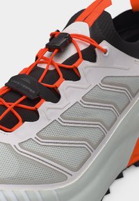 Κοντινή λήψη ενός γκρι και λευκού αθλητικού παπουτσιού με φωτεινά πορτοκαλί ελαστικά κορδόνια στερεωμένα με μαύρο συγκρατητήρα κορδονιών και πλαστικό κουμπωτήρα.