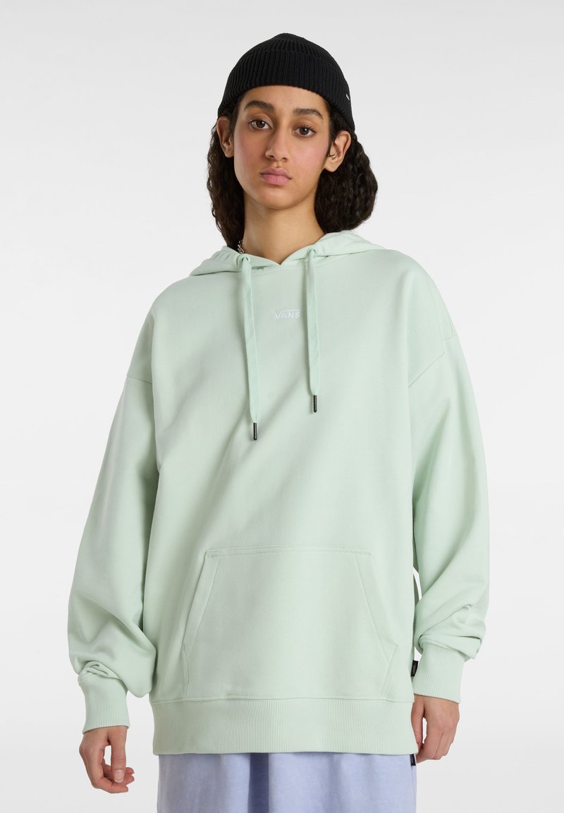 Young person with long curly hair wearing black beanie and oversized pale green Vans hoodie with front pocket on white background.