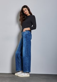 Jeans in denim blu a gamba larga con una texture sbiadita, abbinati a un top nero a coste aderente e sneakers bianche, su uno sfondo neutro.