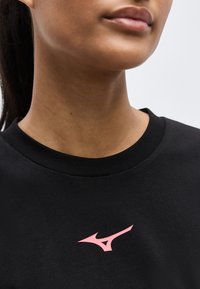 Κοντινό πλάνο ενός ατόμου που φοράει μαύρο μπλουζάκι με στρογγυλή λαιμόκοψη και ένα μικρό ροζ λογότυπο Mizuno στο στήθος.