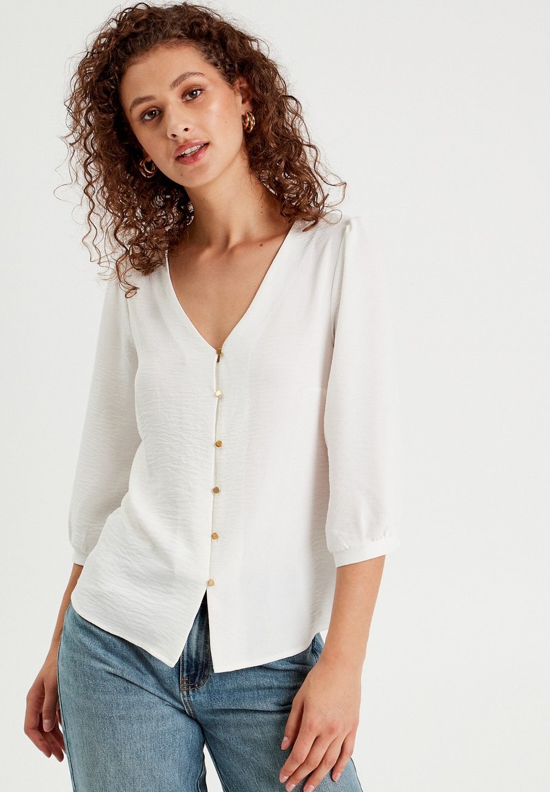 Blouse blanche, légère, avec un décolleté en V, des manches trois-quarts et des boutons dorés sur le devant. Texture lisse avec une coupe décontractée.