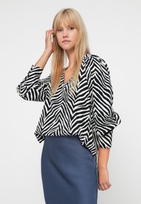 Blouse à rayures zébrées noir et blanc avec un décolleté en V, coupe ample et manches trois-quarts à poignets, associée à une jupe crayon bleu marine.