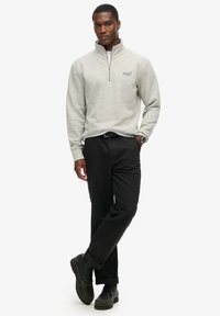 Suéter de media cremallera en gris claro, con una textura suave, combinado con pantalones negros y zapatos negros. Detalles notables incluyen un logo en el pecho.