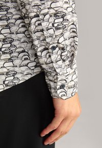 Weißes Hemd mit einem schwarz-grauen wiederkehrenden Muster von Hüten. Verfügt über einen Knopfmanschette mit einem dunklen Knopf. Schwarzer Stoff darunter.