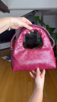 Handen houden een kleine, gestructureerde dieproze handtas met een gebogen bovenhandvat en schouderriem in een lichte binnensetting.