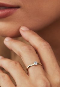 Goldene Verlobungsring mit einem runden, klaren Edelsteineinsatz, getragen an einer Hand mit heller Hautfarbe und natürlichem Nagellack.