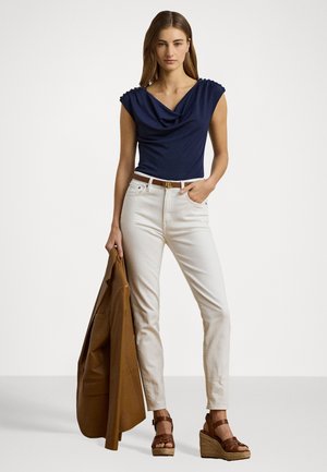 Haut navy drapé avec des manches courtes, associé à un jean blanc ajusté. Modèle tenant un blazer marron, portant des sandales à talons compensés beiges.