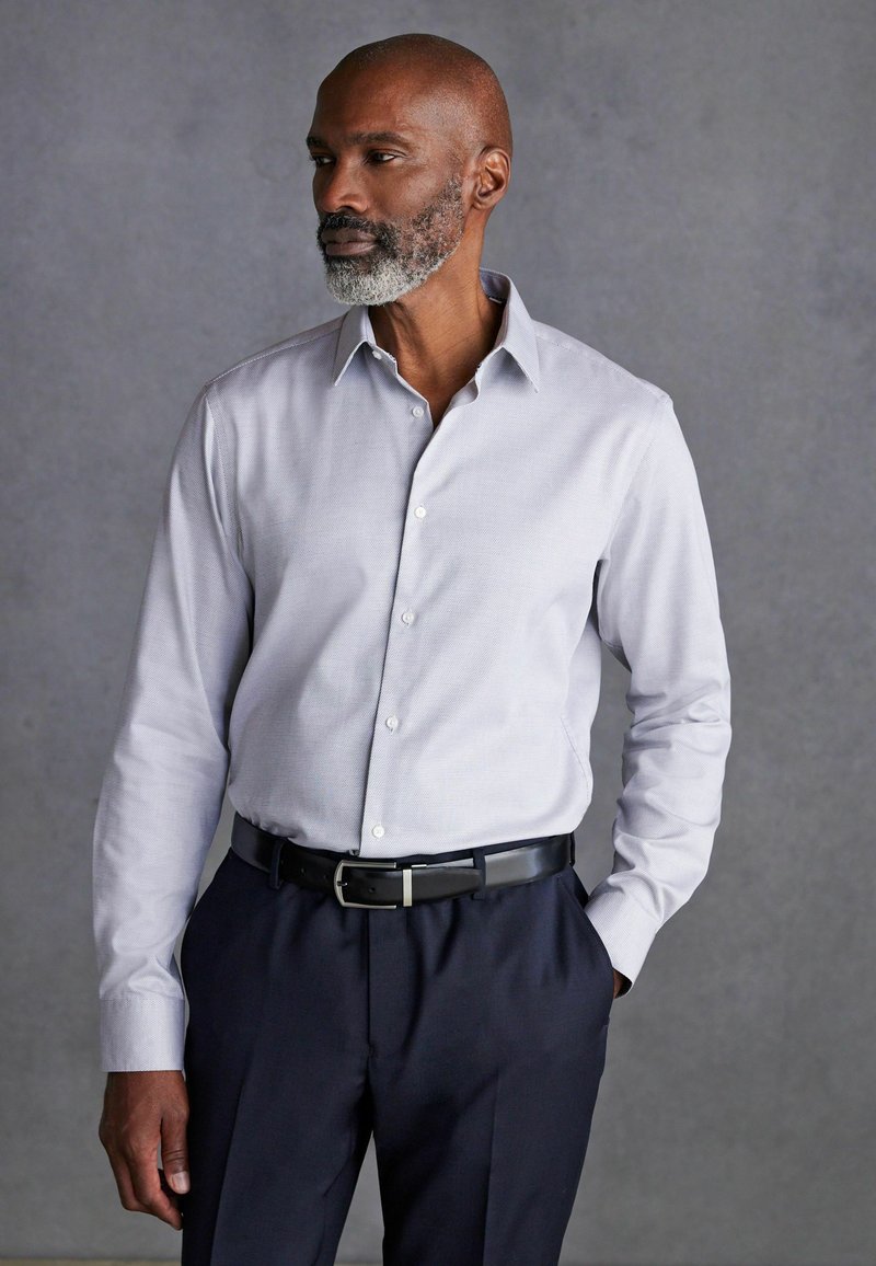 Chemise habillée à manches longues gris clair avec un motif subtil, col boutonné, associée à un pantalon sombre et une ceinture noire avec une boucle en argent.