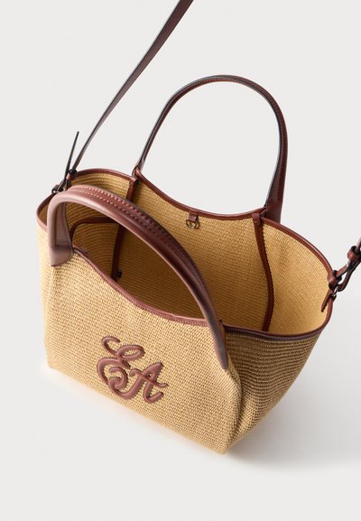 Πλεκτό μπεζ τσάντα tote με καφέ δερμάτινη λεπτομέρεια και λαβές, με πλεγμένα δερμάτινα αρχικά "SA" στο μπροστινό μέρος, ανοιχτό εσωτερικό ορατό.