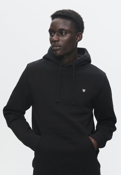 Jeune homme portant un sweat à capuche noir avec une poche avant et un petit logo, regardant sur le côté, les mains dans les poches, fond blanc neutre.